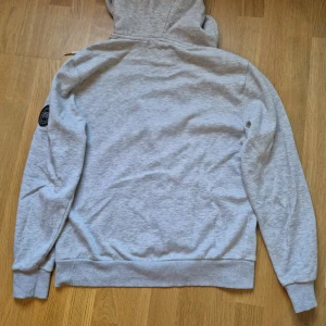 Ljusgrå hoodie från Canada Goose - Snygg ljusgrå hoodie från Canada Goose med dragkedja, huva med snörning och klassisk logga på ärmen. Tillverkad i mjuk bomullsmix, perfekt för chill dagar. Fickor framtill och ribbade muddar för extra komfort.