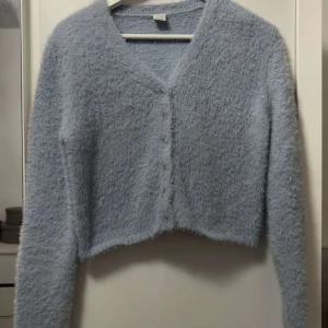 Ljusblå fluffig kofta från Lindex - Supermysig ljusblå kofta från Lindex med fluffig yta och v-ringad hals. Koftan har långa ärmar, knappar framtill och är croppad i modellen. Perfekt för dig som gillar mjuka och gosiga plagg med chill vibe.