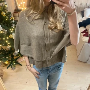 Stickad poncho jacka  - Superfin beige stickad väst från Zara med rund hals och knappar framtill. Modellen är lite oversized och har en cape-liknande form med breda ärmhål och ribbade kanter. Perfekt att slänga över en t-shirt eller skjorta för en chill look.
