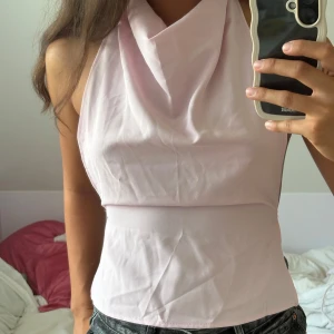 Ljusrosa omlott halterneck topp - Supersöt ljusrosa halterneck topp med snygg draperad halsringning och knyt i ryggen. Toppen är ärmlös och har en omlott-detalj som ger en riktigt trendig vibe. Perfekt till jeans eller kjol för en fräsch look.