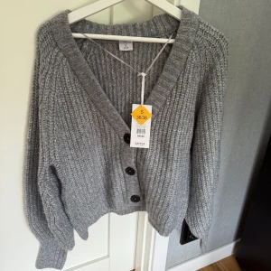 Grå stickad kofta från Stories - Säljer en  grå stickad kofta från Stories med bred ribbad kant och tre svarta knappar framtill. Koftan har en djup v-ringning och långa ärmar med ribbade muddar. Aldrig använd och prislapp kvar
