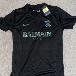 Svart PSG x Balmain fotbollströja Nike - Svart fotbollströja från Nike i samarbete med PSG och Balmain. Tröjan har korta ärmar, rund hals och är tillverkad i lätt funktionsmaterial. Framsidan pryds av holografiska Balmain Paris-tryck och PSG-emblem. Perfekt för dig som vill sticka ut på planen.