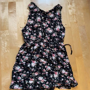 Blommig kortklänning från Catwalk - Supersöt svart kortklänning med blommigt mönster i rosa och vitt från Catwalk on the beach. Klänningen har ärmlös design, knyt i midjan och dekorativa små bollar längs kanterna. Perfekt för sommarens alla tillfällen!