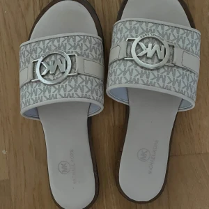 Vita sandaler från Michael Kors - Snygga sandaler från Michael Kors i vitt med MK-logga i silver framtill. Bred rem med mönstrat MK-tryck och metallspänne. Platt sula och öppen tå, perfekt för varma dagar. Stilsäkra och lätta att matcha till sommarens outfits.