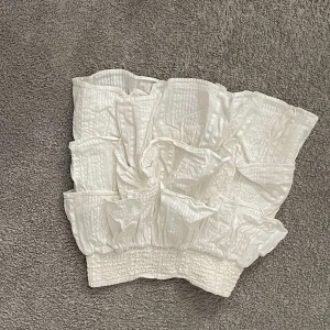 Vit bandeau tubtopp med volanger - Supersöt vit bandeau tubtopp med volangdetaljer och smockad nederkant. Toppen har en lätt glansig yta och struktur i tyget som ger en cool och trendig look. Perfekt att styla till jeans eller kjol för en fräsch sommaroutfit.