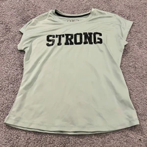 Mintgrön t-shirt från OTP med tryck - Fräsch mintgrön t-shirt från OTP med svart text 'STRONG' på bröstet. T-shirten har rund hals och korta ärmar, perfekt för träning eller chill. Materialet är mjukt och lätt polyester som andas bra. Enkel och stilren design som passar dig som gillar sportiga vibes.
