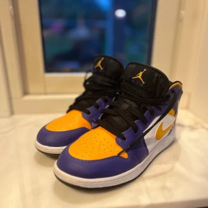Air jordans (Lakers färg tema) - Beskrivning: 	•	Modell: Air Jordan 1 Mid (GS – Grade School storlek) 	•	Storlek: US 6Y (motsvarar ungefär EU 38,5) 	•	Färg: Lila, gult och vitt med svart tunga och svarta skosnören. 	•	Material: Läderöverdel med perforerad tåbox, vadderad plös och krage. 	•	Loggor: 	•	Gult Jumpman-märke och lila “AIR”-text på plösen. 	•	Vit Air Jordan Wings-logga på yttre ankeln. 	•	Gul Nike Swoosh på sidorna. 	•	Yttersula: Svart gummisula med klassiskt Air
