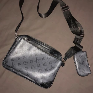 Louis Vuitton axelväska monogram svart/grå - Snygg axelväska från Louis Vuitton i svart och grått med klassiskt monogrammönster. Väskan har justerbar axelrem med logga, dragkedja och en liten avtagbar plånbok. Tillverkad i skinn med silverfärgade detaljer. Perfekt för dig som vill ha en stilren och trendig väska.