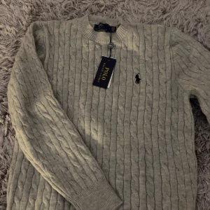  tröja Polo Ralph Lauren - Kabelstickad grå  tröja från Polo Ralph Lauren med klassisk rund halsringning och broderad logga på bröstet. Tröjan har långa ärmar och ribbade muddar. Perfekt för dig som gillar stilren och tidlös design.