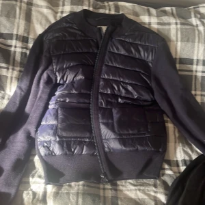 Svart pufferjacka med stickade ärmar - Snygg svart pufferjacka med quiltad framsida och stickade ärmar. Jackan har dragkedja framtill och två stora fickor. Perfekt för dig som vill ha en modern look med både värme och stil.