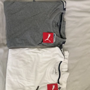 Puma t-shirt grå och vit - Två snygga Puma t-shirts i grått och vitt med klassisk rund hals och korta ärmar. Puma-logga på bröstet och coolt band med Puma-tryck längs axlarna. Perfekt basic med sportig vibe, tillverkad i mjuk bomull för skön känsla.