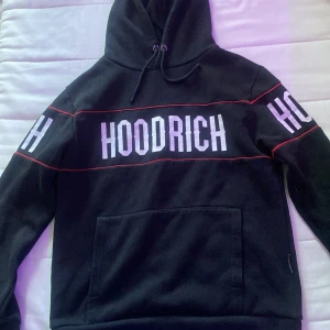 Svart HOODRICH hoodie tracksuit  med röd detalj - Svart hoodie från HOODRICH med stor vit logga på bröstet och ryggen samt på ärmarna. Röd linje går runt överdelen och ärmarna för extra stil. Tröjan har huva med snörning och en stor magficka. Perfekt för dig som gillar streetwear och vill sticka ut.
