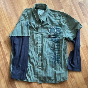 Barn - Grön skjorta med tryck från Garcia Jeans - Tuff grön skjorta från Garcia Jeans med tryck och patchar på bröstet. Skjortan har svarta långa ärmar i lager-på-lager-stil, knappar framtill och råa kanter vid ärmslut. Strl 152