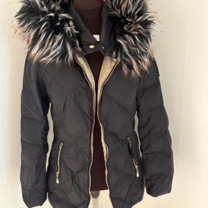 Svart quiltad pufferjacka med fuskpäls - Snygg svart pufferjacka med quiltat mönster och stor fluffig fuskpäls runt huvan. Jackan har dragkedja framtill, två fickor med dragkedjor och coola gulddetaljer. Perfekt för kalla dagar när du vill hålla stilen.