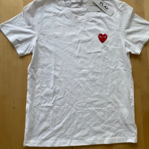 Vit Comme des Garçons Play t-shirt - Snygg vit t-shirt från Comme des Garçons Play med det ikoniska röda hjärtat med ögon broderat på bröstet. Klassisk rund hals och korta ärmar. Tillverkad i mjuk bomull som känns skön mot huden. Perfekt för dig som gillar stilrena plagg med en twist.