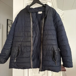 Mörkblå pufferjacka från Zizzi L - Säljer en mörkblå pufferjacka från Zizzi i storlek L. Jackan har horisontella sömmar, två snedställda fickor med dragkedja och hög krage. Perfekt för kyliga dagar och enkel att matcha med olika outfits.
