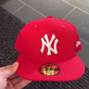 Röd New Era Yankees keps 59FIFTY - Snygg röd New Era 59FIFTY keps med klassiskt New York Yankees-logo broderat i vitt framtill. Platt skärm och broderad New Era-logga på sidan. Kepsen är tillverkad i bomull och har en stilren, sportig look som passar dig som gillar streetwear.