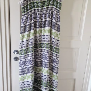 Vit och grön långklänning med mönster - Säljer en boho-inspirerad långklänning med vita, gröna och mörkblå geometriska mönster. Klänningen har resår i midjan och dekorativa fransar längst ner. Perfekt för dig som gillar avslappnad och färgstark stil.
