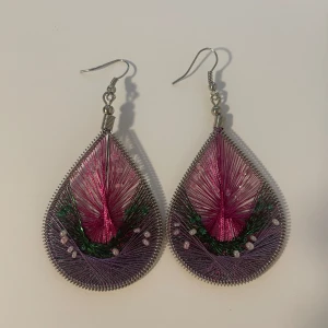 Stora droppformade örhängen i lila och rosa - Unika droppformade örhängen med trådar i lila, rosa och grönt, dekorerade med små pärlor. Metallramen ger en cool kontrast till de färgglada trådarna. Ca 7 cm långa, perfekta för att sticka ut och ge din look en kreativ vibe. Handgjorda, ej av mig.