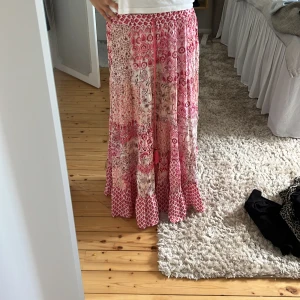 Rosa mönstrad långkjol med snörning - Säljer en flowy långkjol i olika rosa nyanser med bohemiskt mönster och snörning i midjan. Kjolen har volangdetaljer och är perfekt för dig som gillar en avslappnad och färgglad stil.