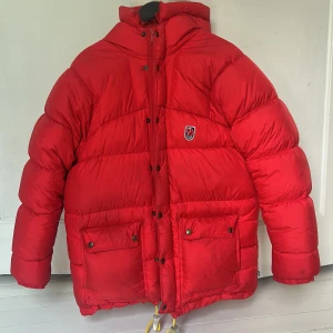 Röd dunjacka från Fjällräven - Säljer nu min dunjacka från Fjällräven med huva och stora fickor framtill. Jackan har klassisk pufferstil, broderad logga på bröstet och dragsko nertill. Perfekt för kalla dagar när du vill hålla värmen och ändå se clean ut. Den har några få mörka fläckar som inte går att få bort men inget man tänker på i verkligheten!
