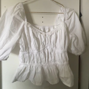 Vit blus med puffärm och spets - Superfin vit blus med puffiga trekvartsärmar och smockad midja. Blusen har v-ringning med söt spetskant och volang nertill. Perfekt för dig som gillar romantisk och luftig stil.