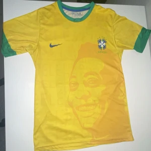 Brasilien Pelé 10 Nike fotbollströja - Gul Brasilien fotbollströja från Nike med gröna detaljer, CBF-emblem och Pelé-tryck med nummer 10 på ryggen. Framsidan har ett stort motiv av Pelé. Klassisk rund hals och korta ärmar. Tillverkad i lätt och ventilerande polyester.