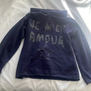 Mörkblå hoodie från Gina Tricot - Säljer en mörkblå hoodie från Gina Tricot med texten 'Je m'en amour' och en stjärna i glittrigt tryck på framsidan. Tröjan har långa ärmar och en bekväm huva. Ärmarna och botten är avklippta då muddarna var slitna!