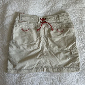 Jeanskjol med detaljer - Snygg beige kortkjol med röda snördetaljer och broderier. Kjolen har en unik design med en mörkblå panel på sidan och praktiska fickor. 