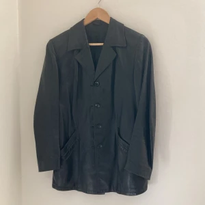 Svart skinnjacka med knappar - Snygg svart skinnjacka med klassisk krage och knäppning framtill. Jackan har långa ärmar och två fickor fram. Storlek ca S