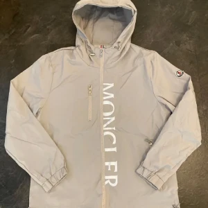 Grå vindjacka från Moncler - Säljer en stilren grå  vindjacka från Moncler med huva och dragkedja. Jackan har Moncler-logga på bröstet och en liten patch på ärmen. Perfekt för blåsiga dagar!