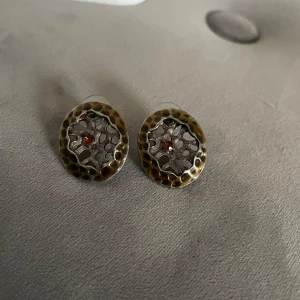 Unika örhängen med mönster - Säljer ett par unika örhängen med ett vackert mönster. Framsidan har en brun och guldaktig ton med en röd detalj i mitten, medan baksidan är i silverfärg. Perfekta för att ge en speciell touch till din stil.