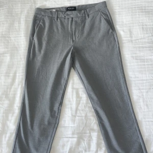 Grå kostymbyxor från Jack & Jones - Snygga grå kostymbyxor från Jack & Jones med slim passform. Byxorna har en klassisk design med hällor för bälte och diskreta bakfickor med knappar. Perfekta för en stilren look.