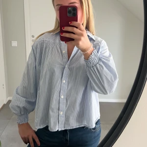 Blåvit randig blus - Säljer en stilren blåvit randig blus med långa ärmar och knappar framtill. Blusen har en lös passform och elastiska ärmslut för extra komfort. Perfekt för en avslappnad men ändå elegant look.🩵