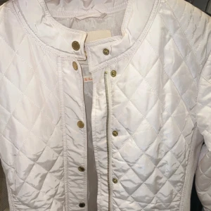 Vit quiltad jacka - Snygg vit quiltad jacka med dragkedja och knappar. Jackan har ett stilrent rutmönster och långa ärmar. Perfekt för vår och höst!