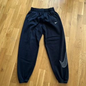Svarta mjukisbyxor/träningsbyxor från Nike - Säljer ett par svarta mjukisbyxor från Nike med elastisk midja och muddar vid bensluten. Byxorna har en liten vit Nike-logga på framsidan och en större logga på benet. Perfekta som en avslappnad eller sportig outfit. Storleken är L i junior och passar alla som är från 165 till 175 centimeter långa. 