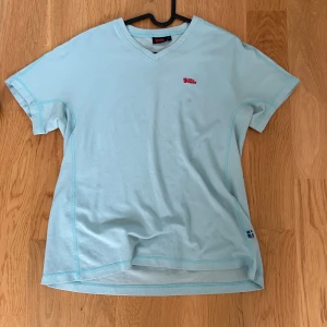 Ljusblå t-shirt från Fjällräven - Säljer en ljusblå t-shirt från Fjällräven med v-ringning och korta ärmar. Den har en liten röd logga på bröstet och är perfekt för en avslappnad stil. T-shirten är tillverkad i ett bekvämt material.