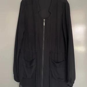 Svart genomskinlig blus med dragkedja - Snygg svart blus med dragkedja framtill och praktiska fickor. Blusen har långa ärmar, see-through /genomskinlig. Storlek 34 från Seppälä. 