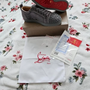 Grå sneakers med nitar från Christian Louboutin - Snygga grå sneakers från Christian Louboutin med ikoniska röda sulor och nitar på tån. Skorna har snörning och är tillverkade i mocka för en lyxig känsla. Perfekta för att ge din outfit en edgy touch.