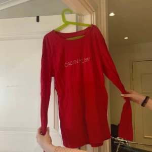 Röd långärmad topp från Calvin Klein - Säljer en snygg röd långärmad topp från Calvin Klein med glittrande logotyp framtill. Perfekt för att ge din outfit en touch av glamour. Toppen är tillverkad i ett mjukt material som känns skönt mot huden.