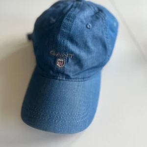 Blå keps från GANT - Snygg blå keps från GANT med broderad logotyp framtill. Kepsen har en justerbar rem baktill med metallspänne för perfekt passform. Perfekt för en avslappnad stil.