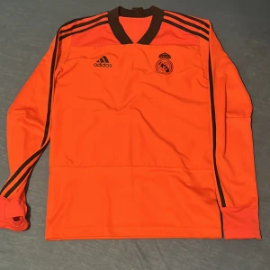 Ljus röd långärmad fotbollströja från Adidas - Säljer en cool ljus röd långärmad fotbollströja från Adidas med svarta detaljer och klubbmärke på bröstet. Tröjan har de klassiska Adidas-ränderna längs ärmarna och en bekväm passform. Perfekt för fotbollsfans som vill sticka ut på planen! Och som hejar på real Madrid det är en 2017 modell! 