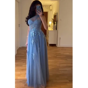 Blå långklänning med blommig spets - Elegant blå långklänning med en axelbandslös design och vackra blommiga spetsdetaljer. Klänningen har en omlottstil och en lätt transparent kjol som ger en drömlik känsla. Perfekt för speciella tillfällen.