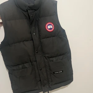 Svart dunväst från Canada Goose - Säljer en svart dunväst från Canada Goose med en klassisk design. Västen har en broderad logotyp på bröstet och praktiska fickor framtill. Perfekt för kyliga dagar.