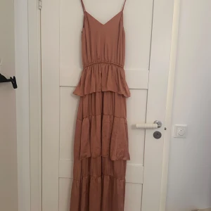 Rosa långklänning med volangdetaljer - Säljer en vacker rosa långklänning med smala axelband och volangdetaljer. Klänningen har en luftig och flödande design, perfekt för varma dagar. Den är tillverkad i ett mjukt och bekvämt material.