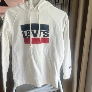 Vit hoodie från Levi's - Säljer en snygg vit hoodie från Levi's med deras ikoniska logga i blått och rött på bröstet. Tröjan har en klassisk känguruficka och justerbar huva med dragsko. Perfekt för en avslappnad stil.