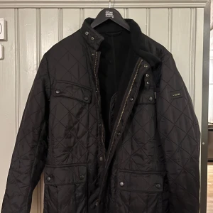 Svart quiltad jacka från Barbour - Säljer en stilren svart quiltad jacka från Barbour International. Jackan har en klassisk design med dragkedja och knappar framtill samt flera praktiska fickor. Perfekt för höst och vår.