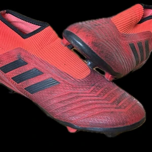 Röda fotbollsskor från Adidas - Snygga röda fotbollsskor från Adidas med svarta detaljer och ikoniska tre ränder på sidan. Skorna har en strumpliknande passform och är designade för att ge bra grepp på planen.