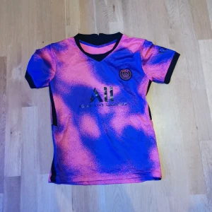 Rosa, blå Messi PSG fotbollströja - Snygg PSG fotbollströja i rosa och blått med korta ärmar. Tröjan har ett coolt mönster och Messi nummer 30 på ryggen. Perfekt för fotbollsfans som vill sticka ut! Trycket nummer 30 är lite knöligt men fotbollströjor med tryck blir så ganska snabbt.