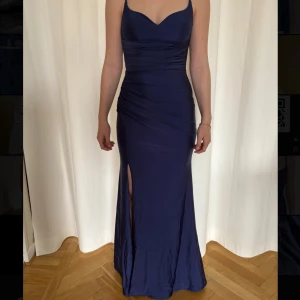 Marinblå balklänning med slits - Elegant marinblå långklänning med smala axelband och en djup v-ringning. Klänningen har en smickrande passform med draperade detaljer och en slits framtill. Perfekt för formella tillfällen. Storleken är mellan XS och S men har en bra stretch i materialet. Endast använd vid 2 tillfällen